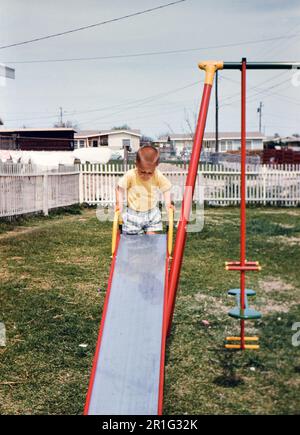 Ein Kleinkind in einem Garten, das sich darauf vorbereitet, eine Rutsche runterzurutschen. 1958 Stockfoto