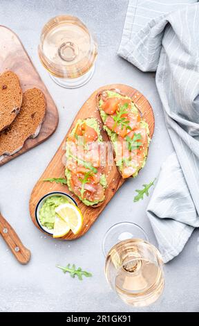 Gourmet-Mittagessen mit Avocado-Lachs-Toasts auf Roggenbrot und Rosenweingläsern, grauer Tischhintergrund, Blick von oben Stockfoto