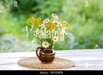 Wunderschöner Strauß von Wildblumen vor dem Hintergrund eines Sommergartens. Blumen in Keramikvase auf Korbmatte auf einem weißen Holztisch. Stockfoto