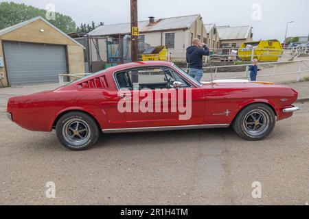 Rayleigh, Großbritannien. Mai 2023. Ein roter Ford Mustang Fastback aus den 1960er Jahren parkte bei einer Autoversammlung in Rayleigh, Essex. Der Muscle Car hat schwarze Räder, verchromte Zierleisten und einen weißen Seitenstreifen. Vor Industriebauten und einem geschlossenen Garagentor stehen Zuschauer und Fotografen in der Szene, die den Geist der britischen Automobilbegeisterung einfangen. Farm Fresh Garage Truck und amerikanische Fahrzeuge werden gezeigt. Penelope Barritt/Alamy Live News Stockfoto