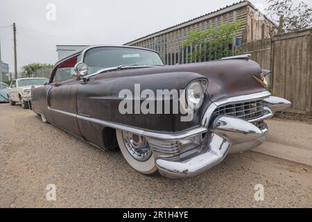 Rayleigh, Großbritannien. Mai 2023. Tiefwinkelansicht eines Vintage Cadillac mit Chromdetails und Whitewall-Reifen, parkt auf einer Schotterstraße in Rayleigh, Essex. Ein zweiter Oldtimer und ein Wellblechgebäude erscheinen im Hintergrund, das an die amerikanische Mitte des Jahrhunderts in britischer Umgebung erinnert. Farm Fresh Garage Truck und amerikanische Fahrzeuge werden gezeigt. Penelope Barritt/Alamy Live News Stockfoto