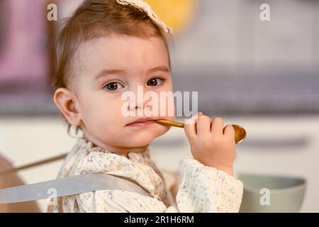 Kleines Mädchen, das in einem Hochstuhl sitzt und mit einem Löffel alleine isst Stockfoto