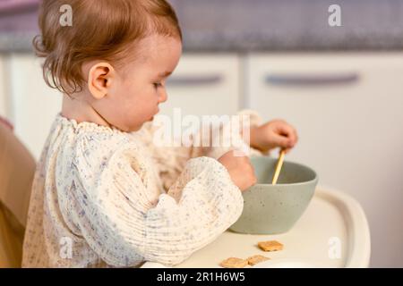 Kleines Mädchen, das in einem Hochstuhl sitzt und mit einem Löffel alleine isst Stockfoto