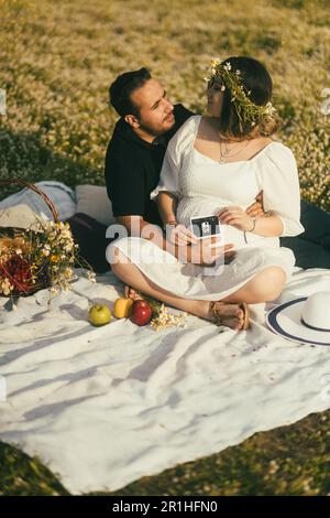 Auf einer weißen Picknickdecke sitzt eine schwangere Frau mit Gänseblümchen-Krone und ihr Mann, die einen fröhlichen Moment teilen, er berührt ihren Bauch, während sie Profi wird Stockfoto