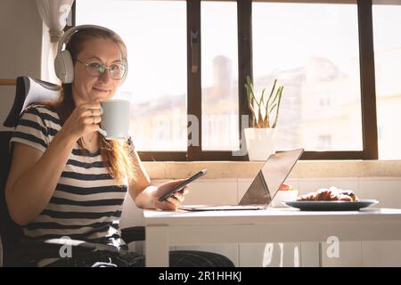Lächelnde Geschäftsfrau mit Kopfhörern, die am Küchentisch sitzt und Kaffee trinkt - und in die Kamera schaut Stockfoto