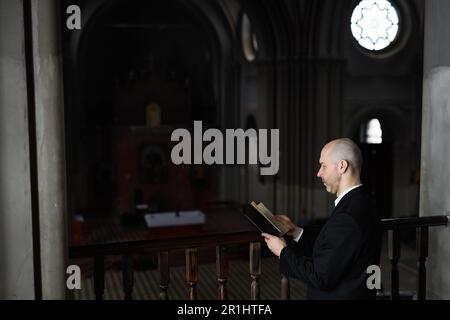 Reifer Priester im schwarzen Anzug, der Bibel liest und betet, während er vor dem Altar in der Kirche steht Stockfoto