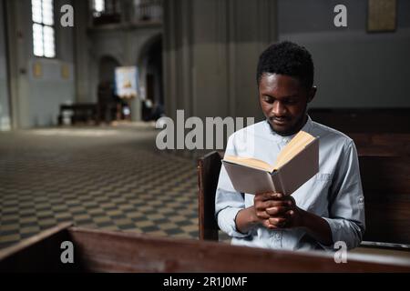 Afroamerikanischer junger Mann, der in der Kirche auf einer Bank sitzt und Gebete liest Stockfoto