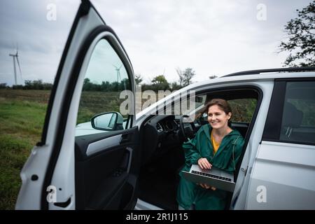 Eine Frau sitzt in einem Auto mit einem Laptop auf einem Hintergrund von Windturbinen. Hochwertiges Foto Stockfoto