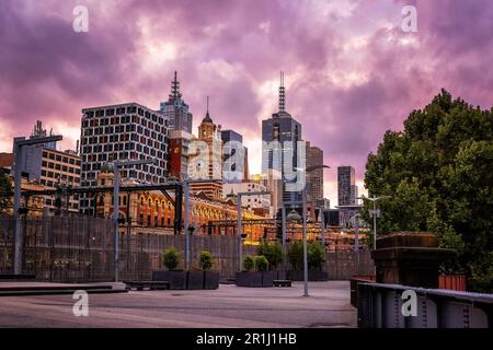 Stadtbild von Melbourne, mit Bahnhof Flinders Street im Vordergrund, in der Abenddämmerung vom Fußweg entlang des Ufers des Yarra River. Leuchtend rosa Su Stockfoto