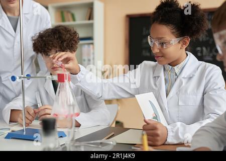 Seitliches Porträt eines lächelnden schwarzen Schulmädchens, das in der Schule Experimente durchführt und Laborkittel trägt Stockfoto