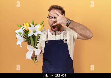 Ein Mann mittleren Alters mit Bartfloristenladen, der Blumen hält, die in den Schock gucken, Gesicht und Augen mit der Hand verhüllen, mit peinlicher Erfahrung durch die Finger schauen Stockfoto