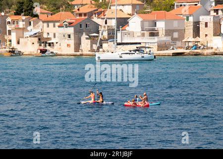 Prvic Luka, Kroatien - 22. Juli 2022: Leute paddeln, während sie auf Paddleboards und Steinhäusern am Dock dahinter sitzen Stockfoto