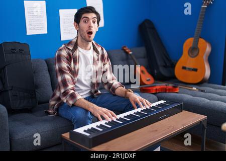 Ein junger lateinamerikanischer Mann, der im Musikstudio Klavier spielt, ängstlich und schockiert vor Überraschungen und erstauntem Gesichtsausdruck, Angst und aufgeregtem Gesicht. Stockfoto