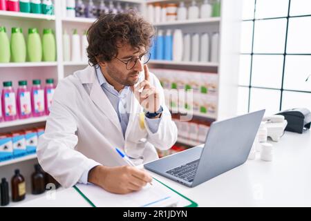 Junger hispanischer Apotheker, der mit dem Laptop in der Apotheke auf Dokumente schreibt Stockfoto