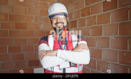 Ein junger lateinischer Baumeister, der selbstbewusst mit gekreuzten Armen auf der Baustelle lächelt Stockfoto