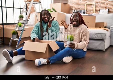 Zwei afrikanische Frauen, die in ein neues Zuhause ziehen, sitzen auf dem Boden und feiern den Sieg mit fröhlichem Lächeln und Gewinnerausdruck mit erhobenen Händen Stockfoto