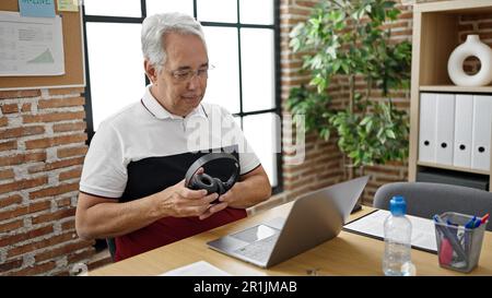 Ein Mann mittleren Alters mit grauhaarigem Büroangestellten, der im Büro einen Laptop mit Kopfhörern benutzt Stockfoto