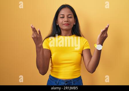 Junge indianerin, die auf gelbem Hintergrund steht, entspannt und mit geschlossenen Augen lächelt und Meditationsgeste mit den Fingern macht. Yoga-Konzept. Stockfoto
