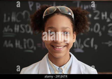 Nahaufnahme eines lächelnden schwarzen Mädchens, das Laborkittel und Schutzbrille im Wissenschaftsunterricht an der Schule trägt und in die Kamera schaut Stockfoto