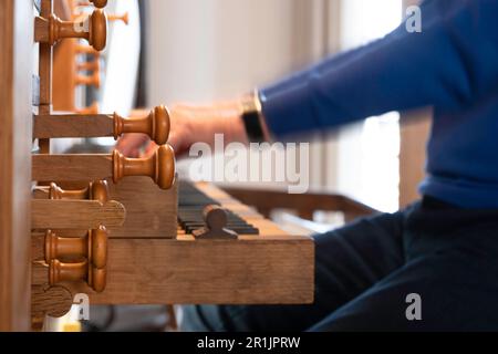 Hände eines Organisten, der eine Kirchenorgel spielt. Von der Seite gesehen, schmale Schärfentiefe, Fokus auf die hölzernen Orgelstopps Stockfoto