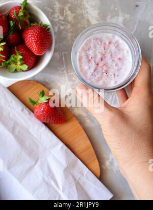 Ansicht von oben: Glasbecher mit ErdbeerSmoothie. Die Hand einer Frau hält ein Glas Smoothie. Auf grauem Hintergrund eine Untertasse mit frischen Erdbeeren. Stockfoto