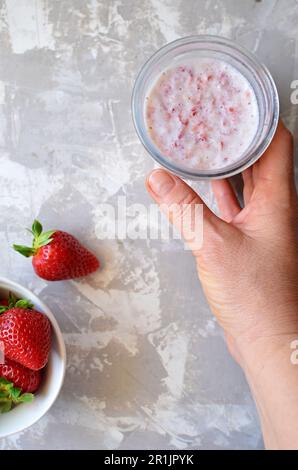 Ansicht von oben: Glasbecher mit ErdbeerSmoothie. Die Hand einer Frau hält ein Glas Smoothie. Auf grauem Hintergrund eine Untertasse mit frischen Erdbeeren. Stockfoto