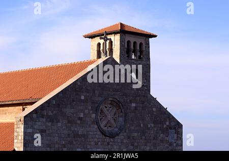 Katholische Kirche im gotischen Stil mit Glockenturm, Kreuz und rotem Dach. Bäume an der Straße und Autos. Tempel am Meer, Mandraki-Platz Stockfoto