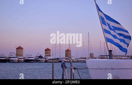 Drei Steinmühlen mit roten Dächern am Ufer. Im Vordergrund befindet sich ein weißes Schiff mit einer winkenden griechischen Flagge. Stockfoto