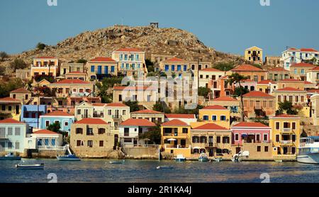 Farbenfrohe Häuser an den Hängen eines Hügels im Hafen der Insel Halki in Griechenland. Kleine Fischerboote liegen vor der Küste im türkisfarbenen Wasser vor. Stockfoto