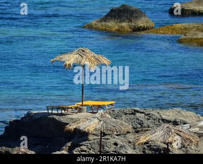 Felsstrand mit Sonnenliegen und Sonnenschirmen in der Nähe des Meeres, auf der Insel. Die Sonnenliegen sind gelb und blau. Strohschirme neben den Sonnenliegen Stockfoto