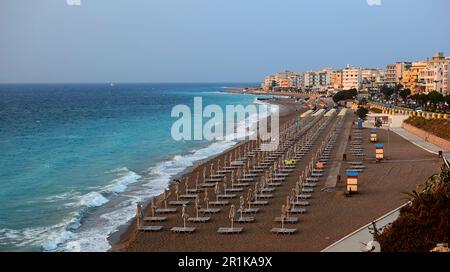 Meeresküste mit blauen Sturmwellen. Entlang der Küste gibt es mehrstöckige Gebäude, am Strand gibt es Reihen von Sonnenliegen mit Sonnenschirmen. Stockfoto