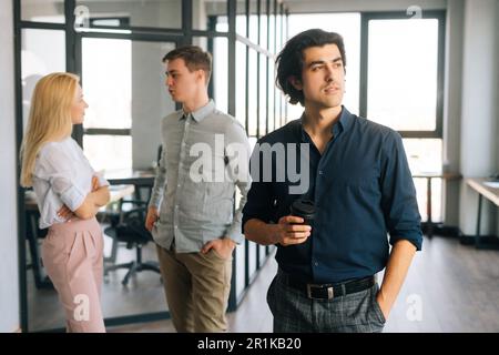 Porträt eines erfolgreichen Startup-Geschäftsmanns in legerer Kleidung, der eine Kaffeetasse zum Mitnehmen in der Hand hält, im Büro steht und selbstbewusst wegschaut. Stockfoto