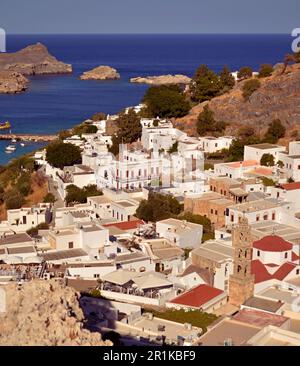 Panoramablick auf das griechische Dorf Lindos mit weißen Häusern und grünen Bäumen am Fuße des Hügels. Felsen und Touristenschiffe sind in der Bucht zu sehen. Stockfoto