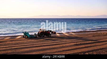 Traktor mit Anhänger zum Reinigen des Strandes von Winterschutt. Der Traktor arbeitet an der Küste, in der Nähe der Wellen. Der Traktor legt Sand auf den Strand. Stockfoto