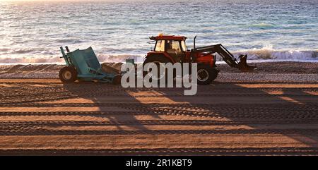 Traktor mit Anhänger zum Reinigen des Strandes von Winterschutt. Der Traktor arbeitet an der Küste, in der Nähe der Wellen. Der Traktor legt Sand auf den Strand. Stockfoto