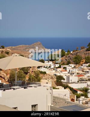 Panoramablick auf das griechische Dorf Lindos mit weißen Häusern und grünen Bäumen am Fuße des Hügels. Felsen und Touristenschiffe sind in der Bucht zu sehen. Stockfoto