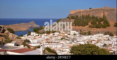 Panoramablick auf das griechische Dorf Lindos mit weißen Häusern und grünen Bäumen am Fuße des Hügels. Felsen und Touristenschiffe sind in der Bucht zu sehen. Stockfoto