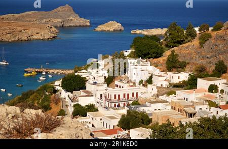 Panoramablick auf das griechische Dorf Lindos mit weißen Häusern und grünen Bäumen am Fuße des Hügels. Felsen und Touristenschiffe sind in der Bucht zu sehen. Stockfoto