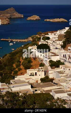 Panoramablick auf das griechische Dorf Lindos mit weißen Häusern und grünen Bäumen am Fuße des Hügels. Felsen und Touristenschiffe sind in der Bucht zu sehen. Stockfoto