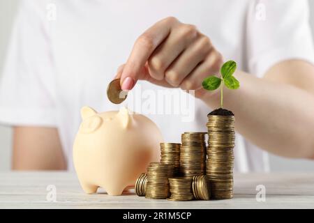 Eine Frau, die Münze in die Schweinebank in der Nähe des Stapels steckt, mit grünem Sprossen am weißen Tisch, Nahaufnahme. Investitionskonzept Stockfoto