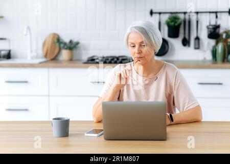 Rätselhafte weißhaarige Dame benutzt einen Laptop, um online zu arbeiten oder im Internet nach Informationen zu suchen, sitzt in der Küche am Schreibtisch, sieht verwirrt auf dem Bildschirm aus, denkt über Informationen nach, Nachrichten Stockfoto
