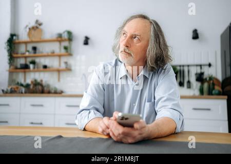 Nachdenklicher, reifer, weißer, grauhaariger Mann mit Bart, sitzt zu Hause in der Küche am Tisch, benutzt sein Smartphone, surft in sozialen Medien, liest Nachrichten, Nachrichten, Er sieht mit Bedacht weg Stockfoto