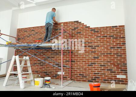 Professioneller Baumeister, der dekorative Ziegel an die Wand im Raum klebt. Installationsvorgang für Fliesen Stockfoto