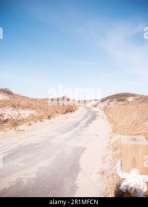 Ein gepflasterter Pfad, der sich durch die Sanddünen neben dem Meer zieht. Der Weg ist mit fließendem Sand bedeckt. Es ist ein warmer Tag mit hellem Sonnenschein Stockfoto