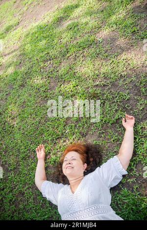 Reife hispanische Frau, die auf dem Gras liegt, mit ihren Armen in einer entspannten Position lächelt und einen Moment der Ruhe genießt. Stockfoto