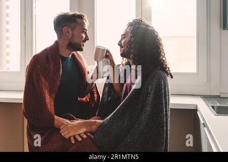 Ein schwules Paar sitzt in der Küche am Fenster, hält Hände und schaut sich in die Augen, ist in eine gemütliche Decke eingewickelt und genießt dabei ein heißes Getränk Stockfoto