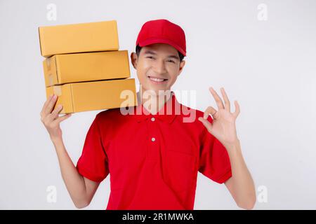 Junger asiatischer Mann in Uniform Rot und Mütze Stehkiste und Geste ok isolierter weißer Hintergrund, Mitarbeiter hält Fracht, Kurier und Zustellung Stockfoto