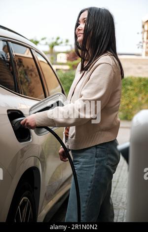 Junge zentralasiatische Frau, die ein Ladekabel anschließt, um ihr Elektroauto aufzuladen. Nachhaltiges und wirtschaftliches Verkehrskonzept. Stockfoto