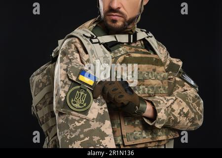 Soldat mit ukrainischer Flagge und Dreizack auf Militäruniform vor schwarzem Hintergrund, Nahaufnahme Stockfoto