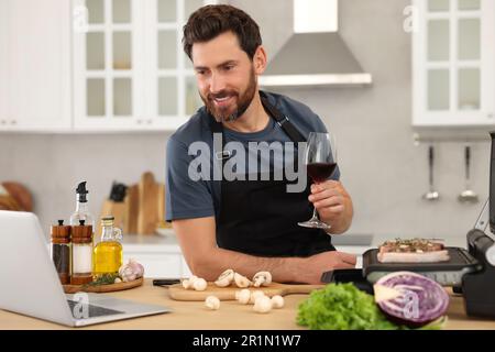 Mann mit einem Glas Wein beim Abendessen, während er sich den Online-Kochkurs mit einem Laptop in der Küche anschaute Stockfoto